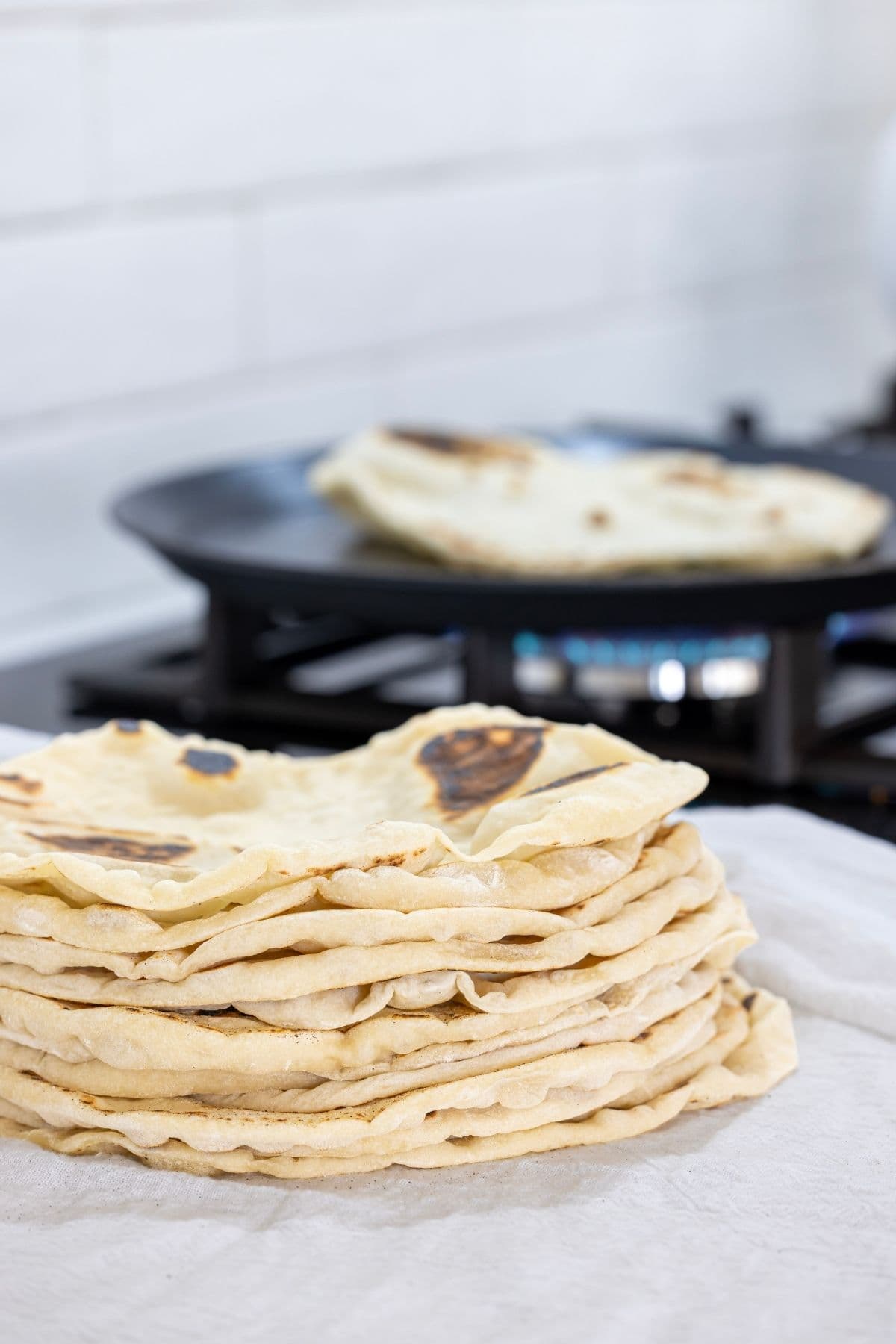 A stack of freshly cooked sourdough discard tortillas stacked on a white kitchen towel. You can see a large open pan in the background with a tortilla being cooked over a blue gas flame.