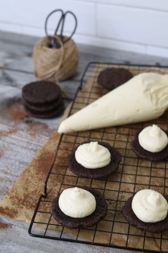 Sourdough chocolate sandwich cookies sitting on a black wire cooling rack. They have been piped with a vanilla buttercream frosting.