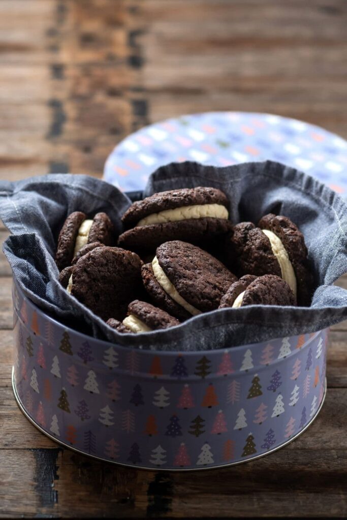 A pale blue and lilac tin lined with a blue dish towel. Inside there are a pile of sourdough chocolate sandwich cookies filled with vanilla buttercream.