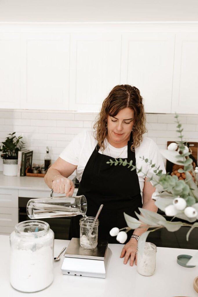 Kate is wearing a black apron and feeding a sourdough starter using a scale. She is pouring water from a glass jug into a 460ml mason jar. There is also a large glass canister of flour on the counter top.