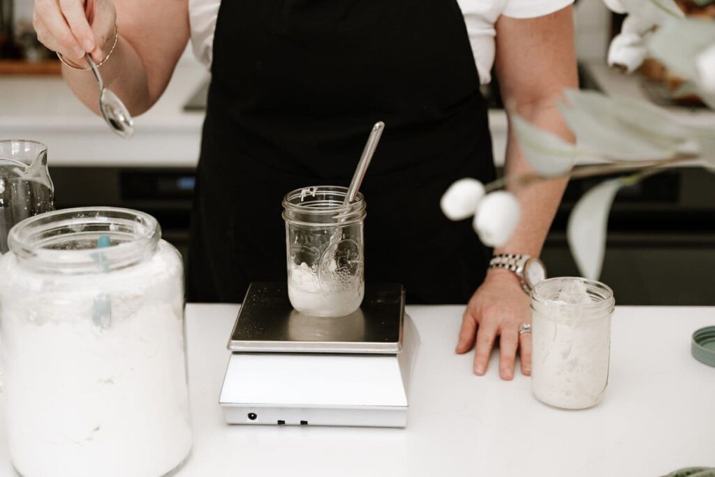 A glass jar sitting on a kitchen scale. You can see someone feeding the sourdough starter with flour and water.