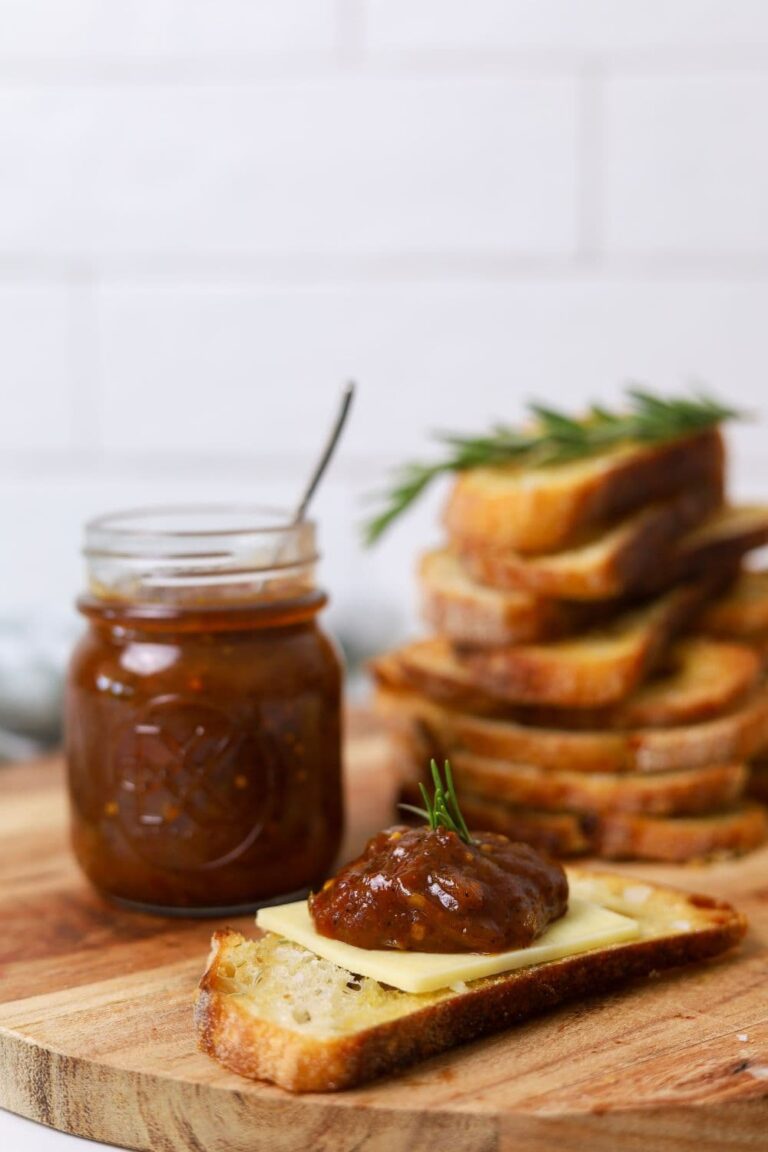 A jar of chilli tomato relish sitting next to a stack of sourdough crostini. One of the crostini has been loaded with cheddar cheese and a big dollop of chilli tomato relish.