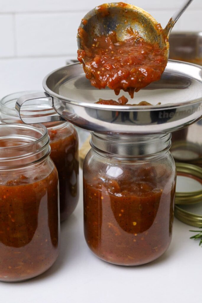 A photo showing the chilli tomato relish being poured into glass jars using a stainless steel jar funnel and ladle.