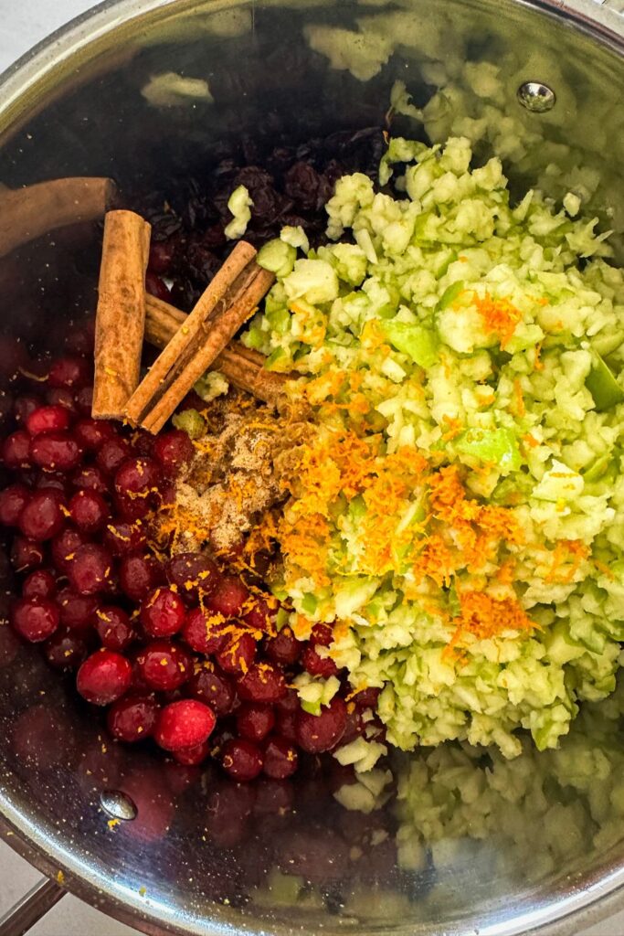 All of the ingredients for making Christmas chutney arranged in a stainless steel saucepan. You can see all the colors of the ingredients layered in the pot.