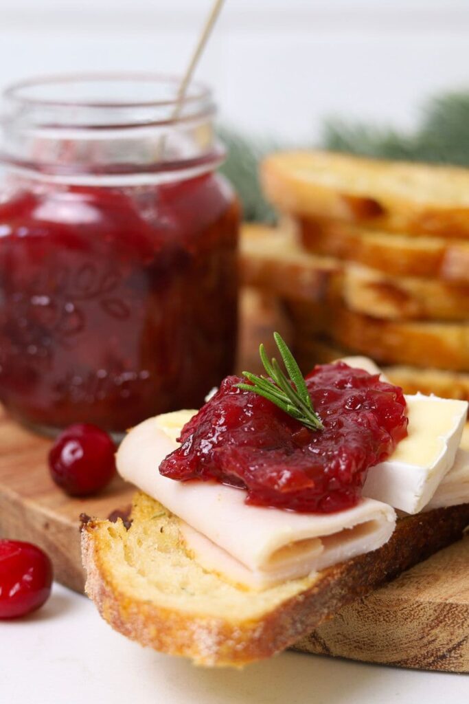 Christmas chutney served on a sourdough crostini topped with turkey and Brie. You can see a jar of Christmas chutney in the background, along with a stack of sourdough crostini.