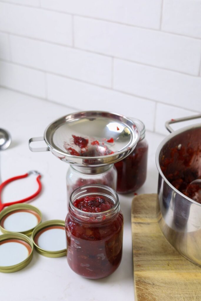 A 500ml glass jar filled with Christmas chutney. You can also see other equipment in the photo including a stainless steel jar funnel and red jar tongs.