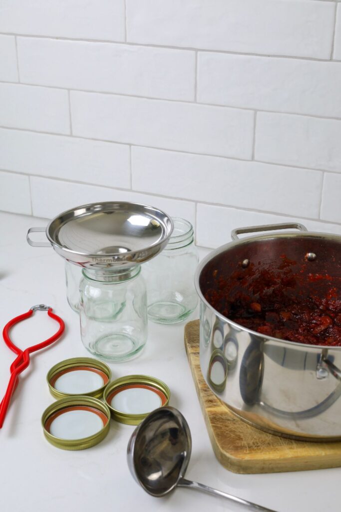 Equipment necessary to make Christmas chutney. In this photo you can see red jar tongs, large stainless steel pot, stainless steel ladle and jar funnel, as well as some 500ml glass jar.