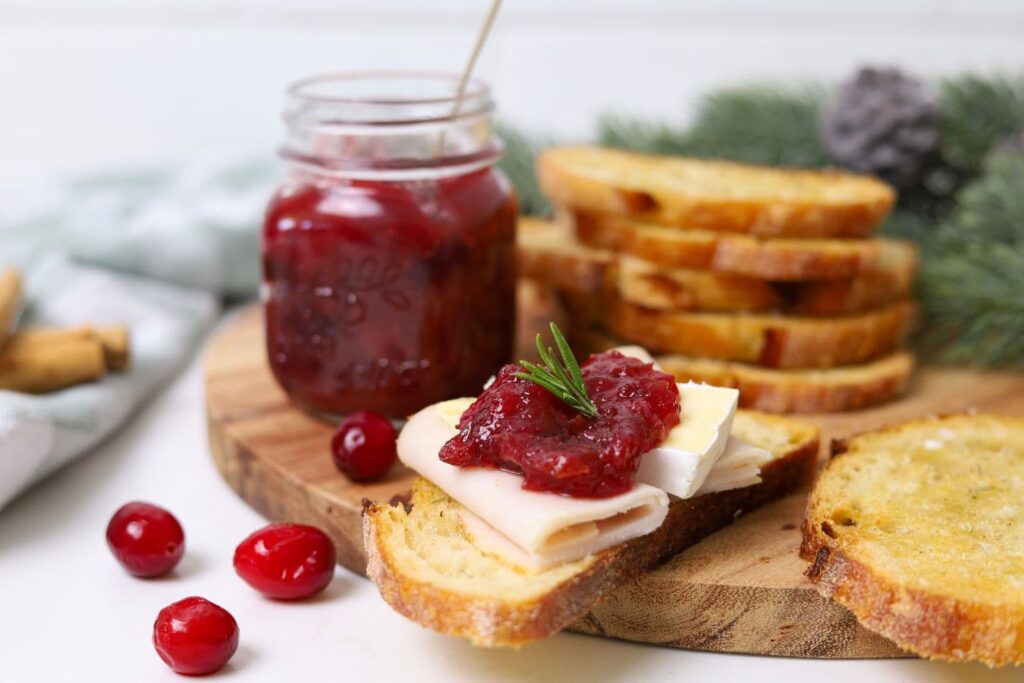 A jar of Christmas chutney sitting next to a pile of sourdough crostini. There is a crostini topped with turkey and chutney.