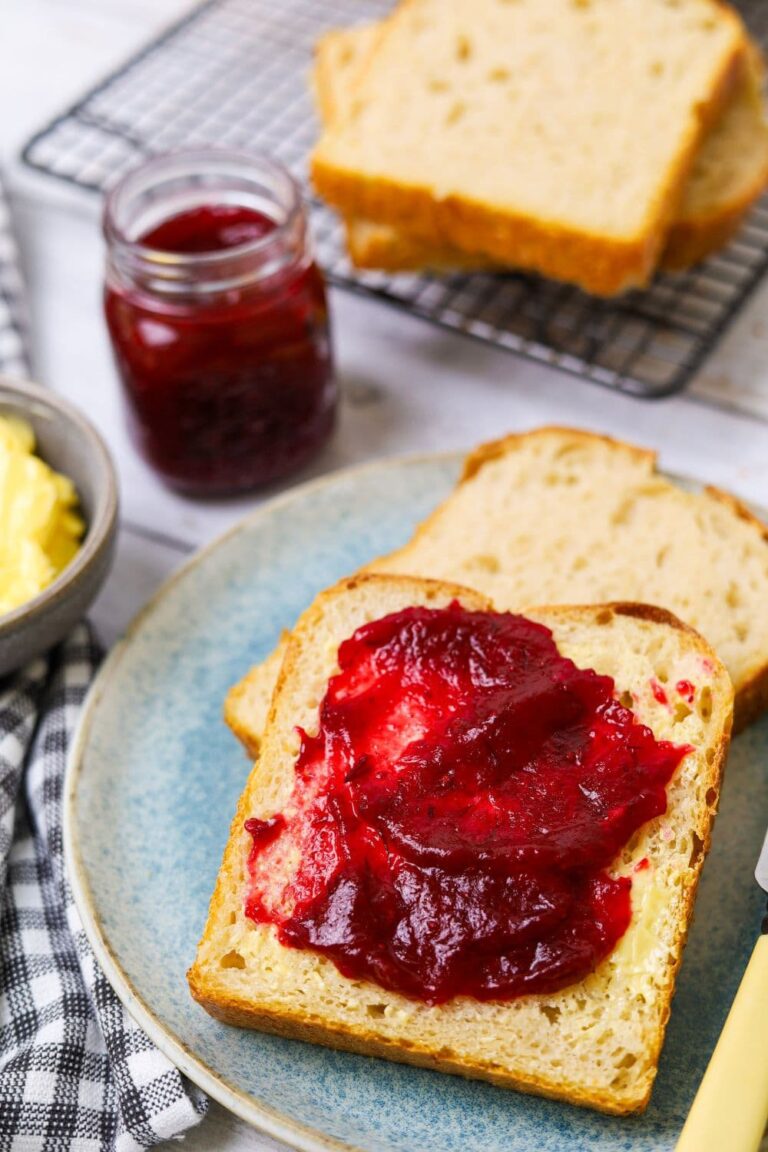 A slice of sourdough bread spread with cranberry orange jam. You can see a small jar of cranberry orange jam in the background of the photo.