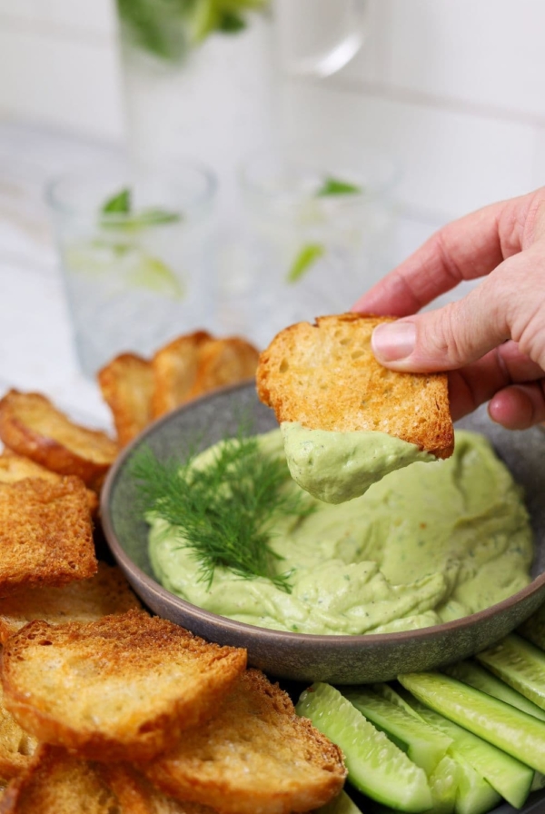 A sourdough crostini being dipped into a grey bowl of bright green smooth avocado dip garnished with fresh dill.