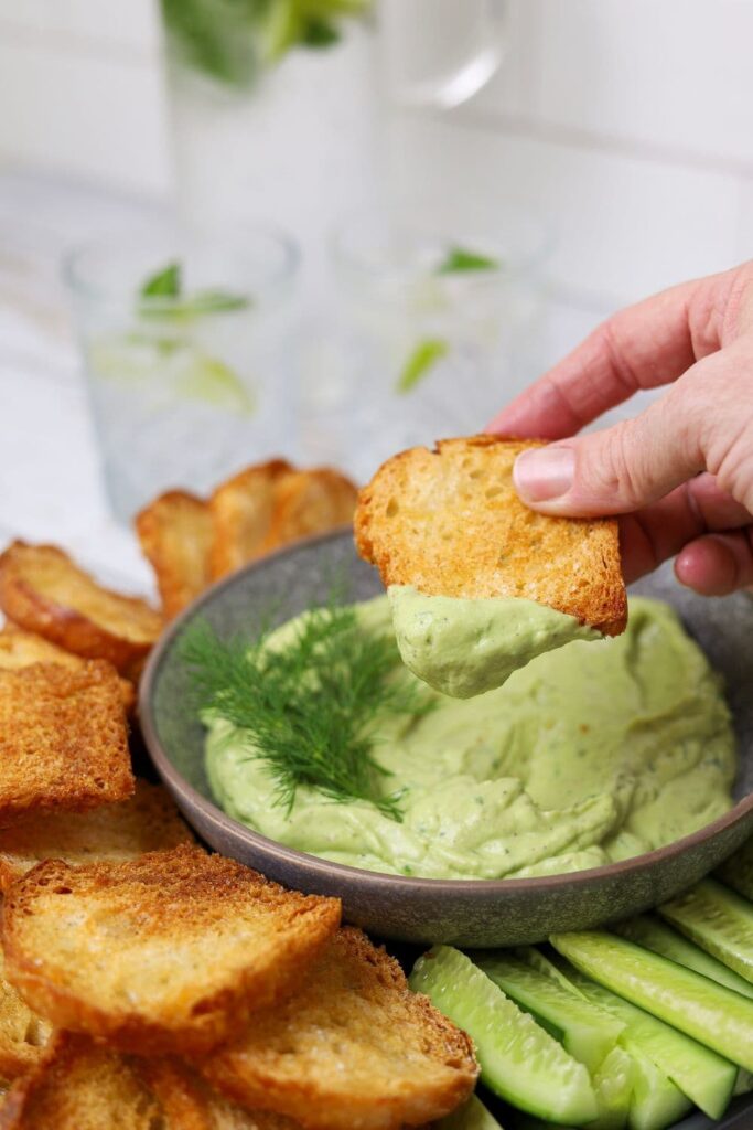 A sourdough crostini being dipped into a grey bowl of bright green smooth avocado dip garnished with fresh dill.