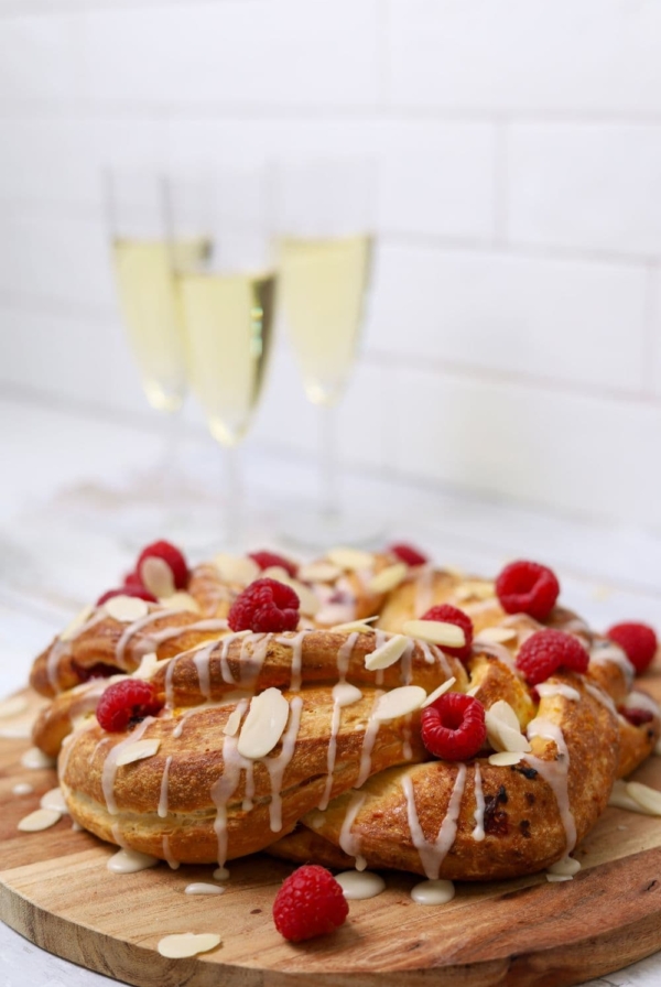 A sourdough brioche braided wreath drizzled in orange glaze and topped with fresh raspberries and flaked almonds. You can see 3 glasses of champagne in the background, evoking a festive ocassion.
