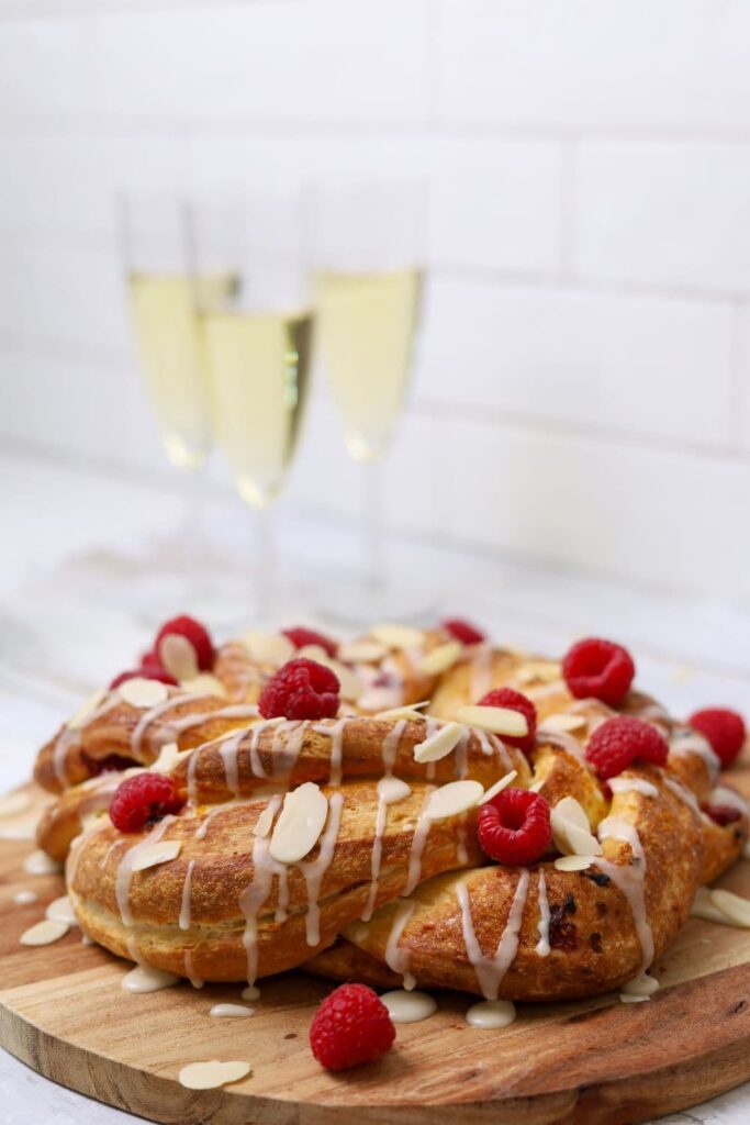 A sourdough brioche braided wreath drizzled in orange glaze and topped with fresh raspberries and flaked almonds. You can see 3 glasses of champagne in the background, evoking a festive ocassion.