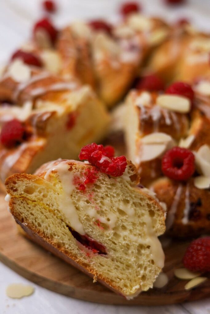 A close up photo of the sourdough brioche braided wreath with a slice cut out and showing the crumb. The wreath is decorated with raspberries and flaked almonds.