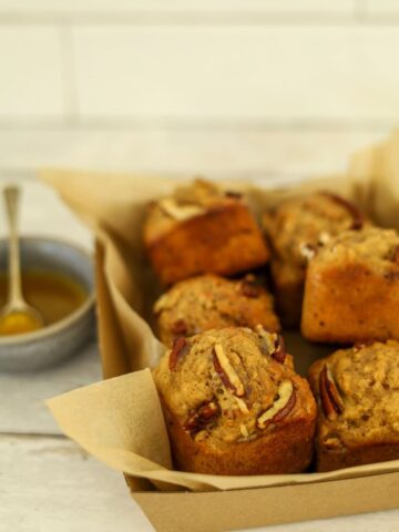 A tray of maple pecan sourdough muffins baked in a square muffin pan. You can see a bowl of maple butter syrup to the left that has been drizzled over the warm muffins.