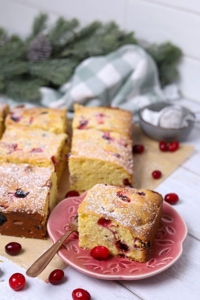 A piece of sourdough cranberry cake sitting on a small pink plate. You can see the rest of the cranberry cake in the background of the photo with some festive decorations.