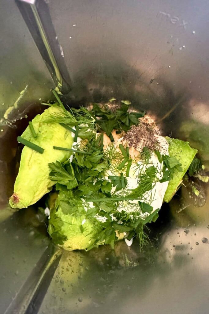 Looking down into a food processor and you can see fresh avocado, yogurt, fresh herbs and all the ingredients ready to be made into smooth avocado dip.