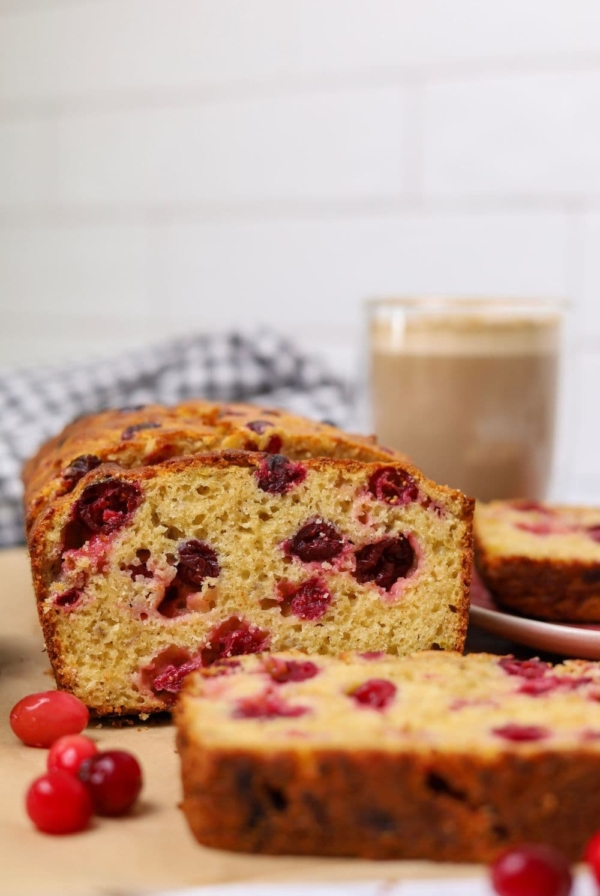 A close up of a slice of sourdough cranberry banana bread. You can see the red cranberries in the cake batter. There is also a cup of coffee in the background.