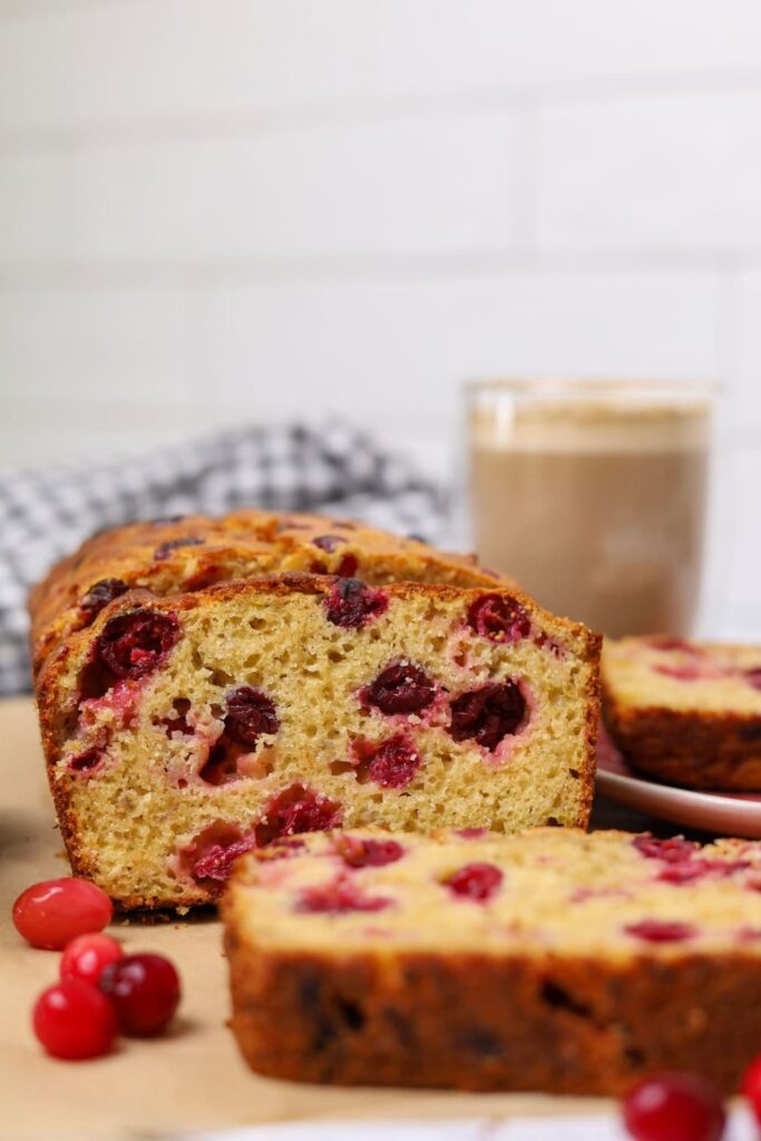 A close up of a slice of sourdough cranberry banana bread. You can see the red cranberries in the cake batter. There is also a cup of coffee in the background.