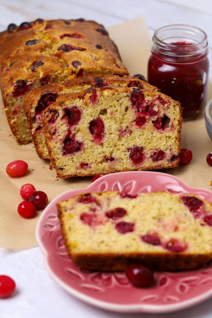 A loaf of sourdough cranberry banana bread that has been sliced up. There is a slice sitting on a small pink plate and some frozen cranberries decorating the scene.