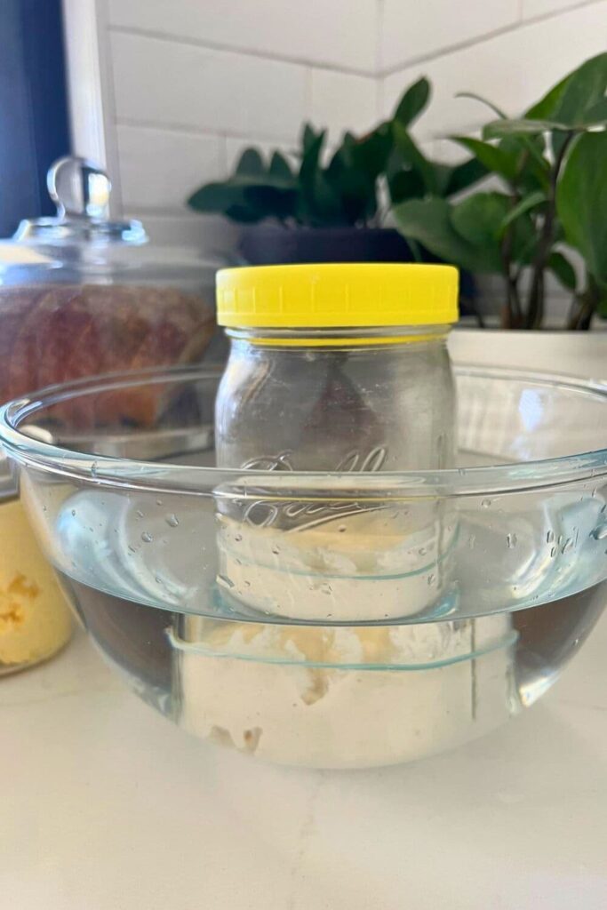 A mason jar with a yellow plastic lid placed in a bowl of warm water. 