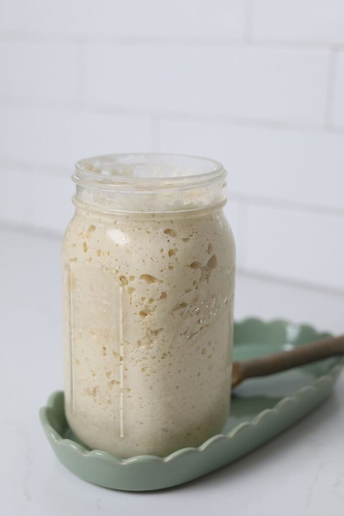 A jar of sourdough starter sitting in a green ceramic tray. The jar of starter is very bubbly and active.