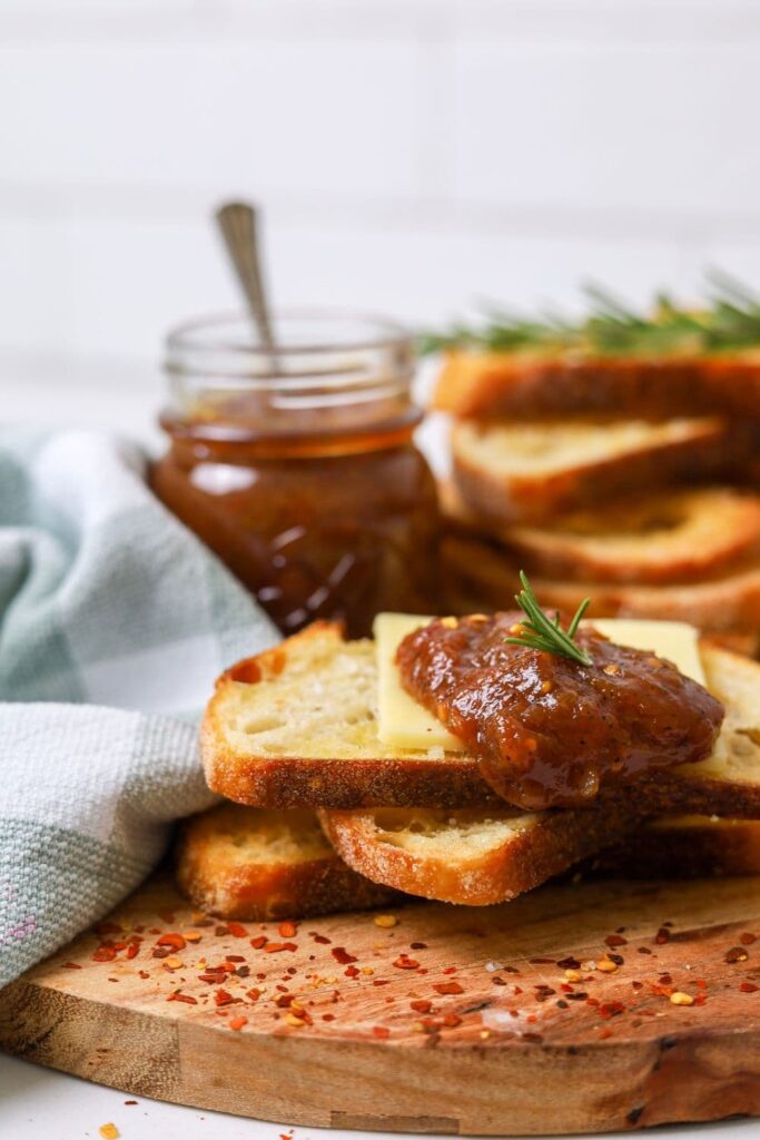 A jar of chilli tomato relish sitting next to a stack of sourdough crostini. One of the crostini has been loaded with cheddar cheese and a big dollop of chilli tomato relish.