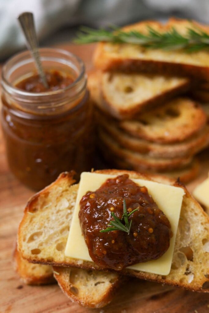 A jar of chilli tomato relish shown sitting alongside a stack of sourdough crostini. One of the crostini has been loaded up with a slice of cheese and dollop of spicy tomato relish.