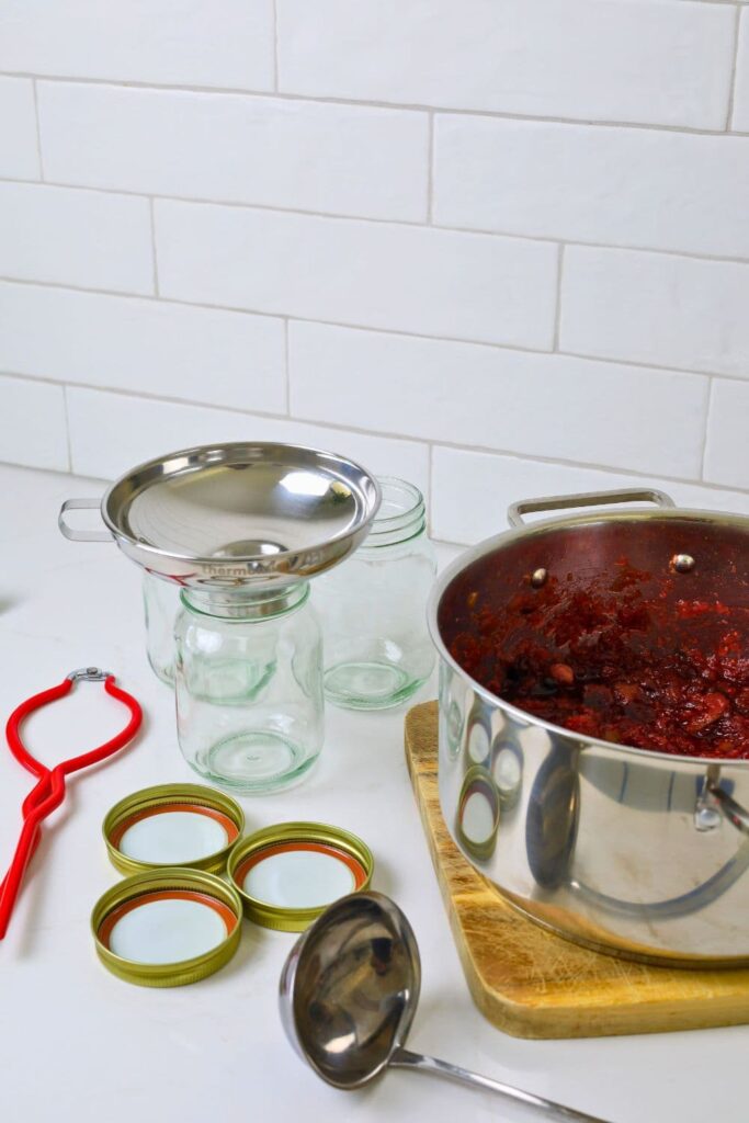 Equipment necessary to make tomato chutney. In this photo you can see red jar tongs, glass jars with gold lids, stainless steel ladle and jar funnel and a stainless steel pot.