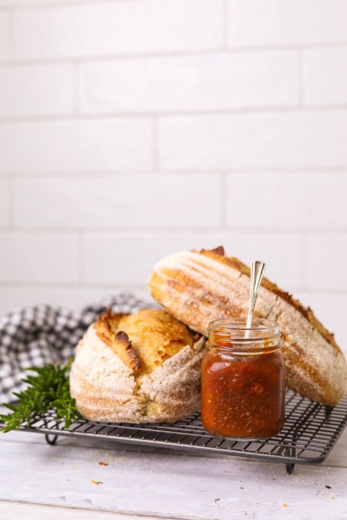 Two loaves of sourdough bread sitting on a wire cooling rack. There is a jar of tomato chutney in front of the loaves of sourdough bread.