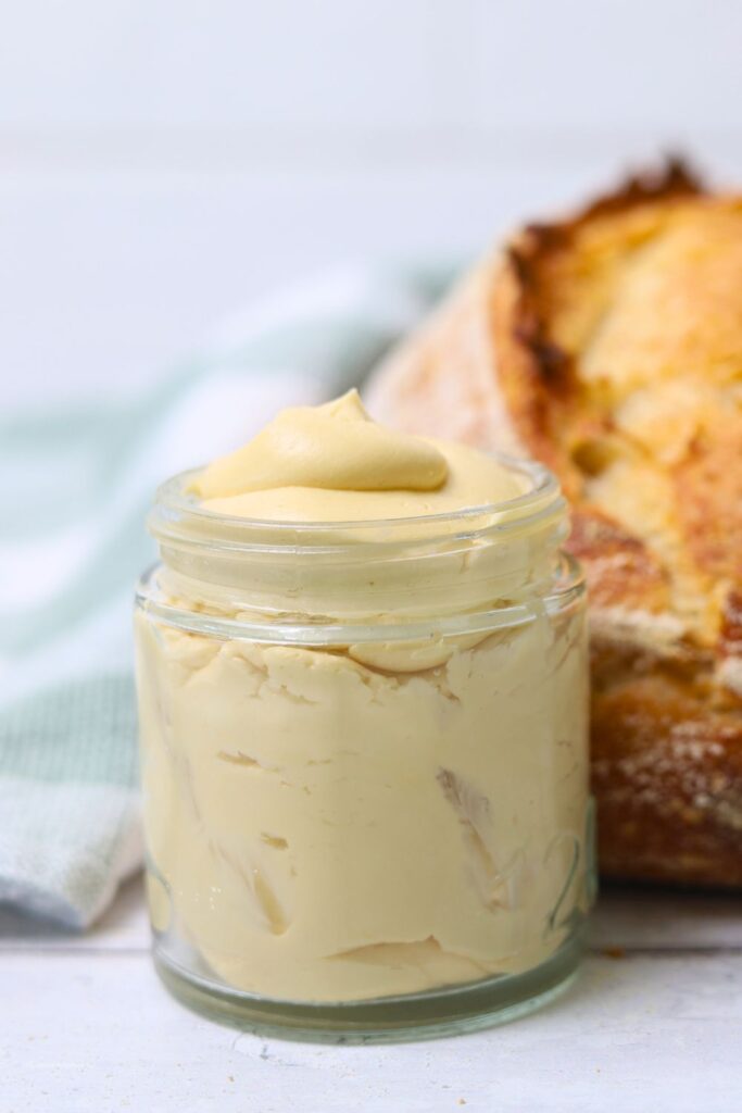 A small glass jar of whipped maple butter sitting in front of a loaf of sourdough bread.