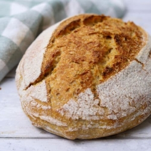 Loaf of whole wheat sourdough bread sitting next to a green and white checked dish towel. Recipe feature image.