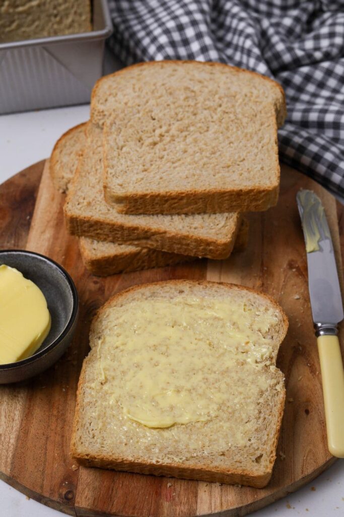 Slices of whole wheat sourdough bread sitting on a round wooden board. The slice at the front has been buttered. 