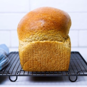 A close up of a loaf of whole wheat sourdough sandwich bread sitting on a wire cooling rack. You can see a light blue dish towel in the background of the image.