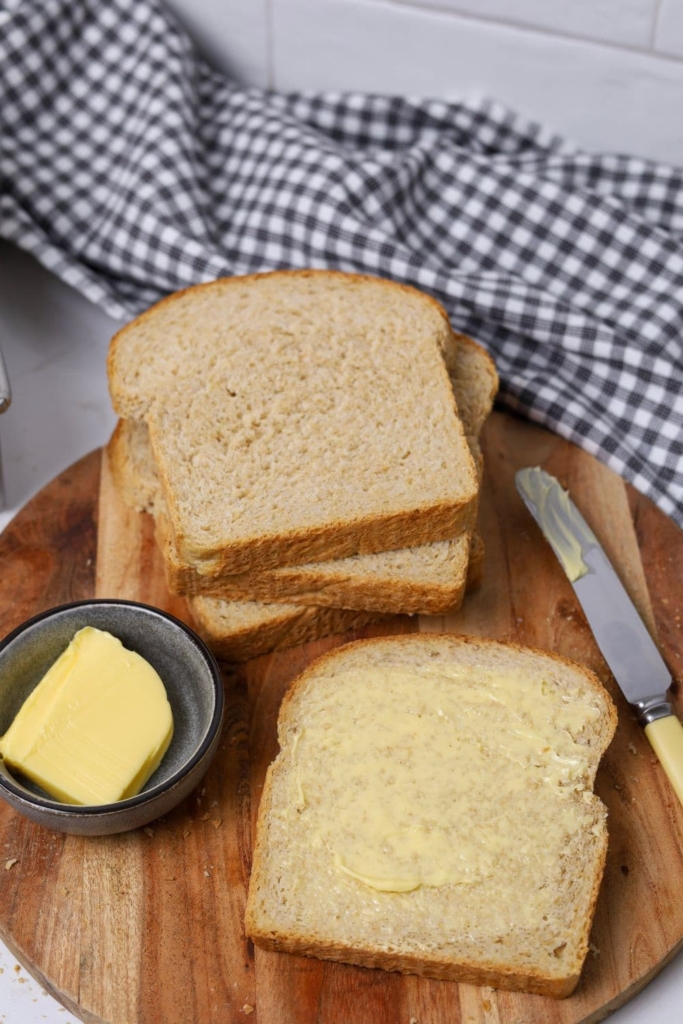 Slices of whole wheat sourdough sandwich loaf sitting on a wooden board with a small bowl of butter.