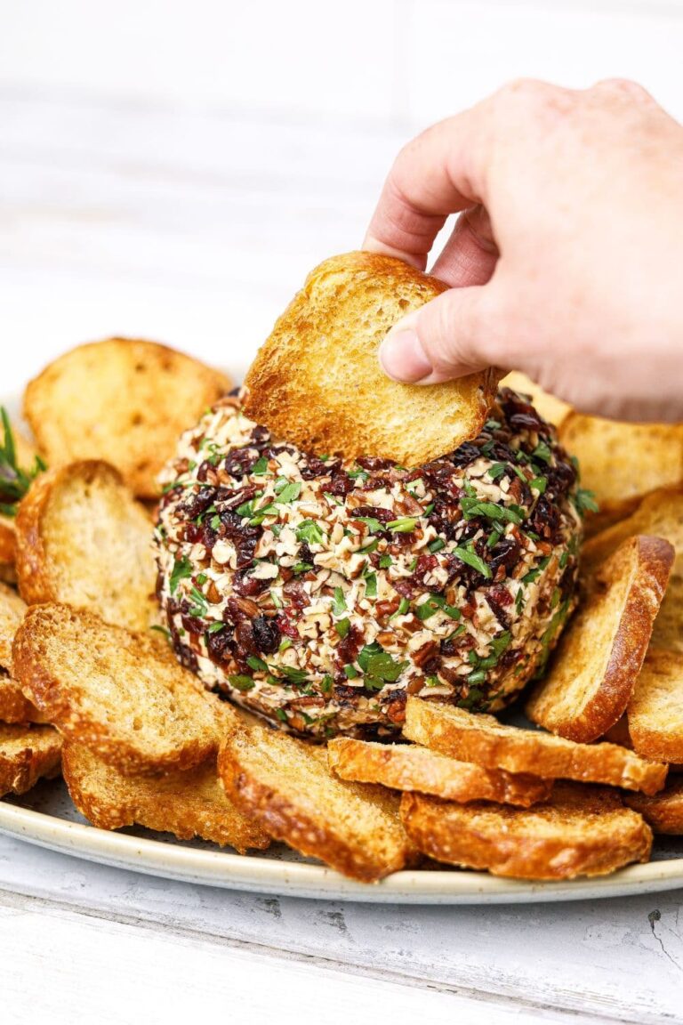 A cranberry pecan cream cheese ball appetiser served with sourdough crostini. There is a crostini being dipped into the top of the cream cheese appetizer.