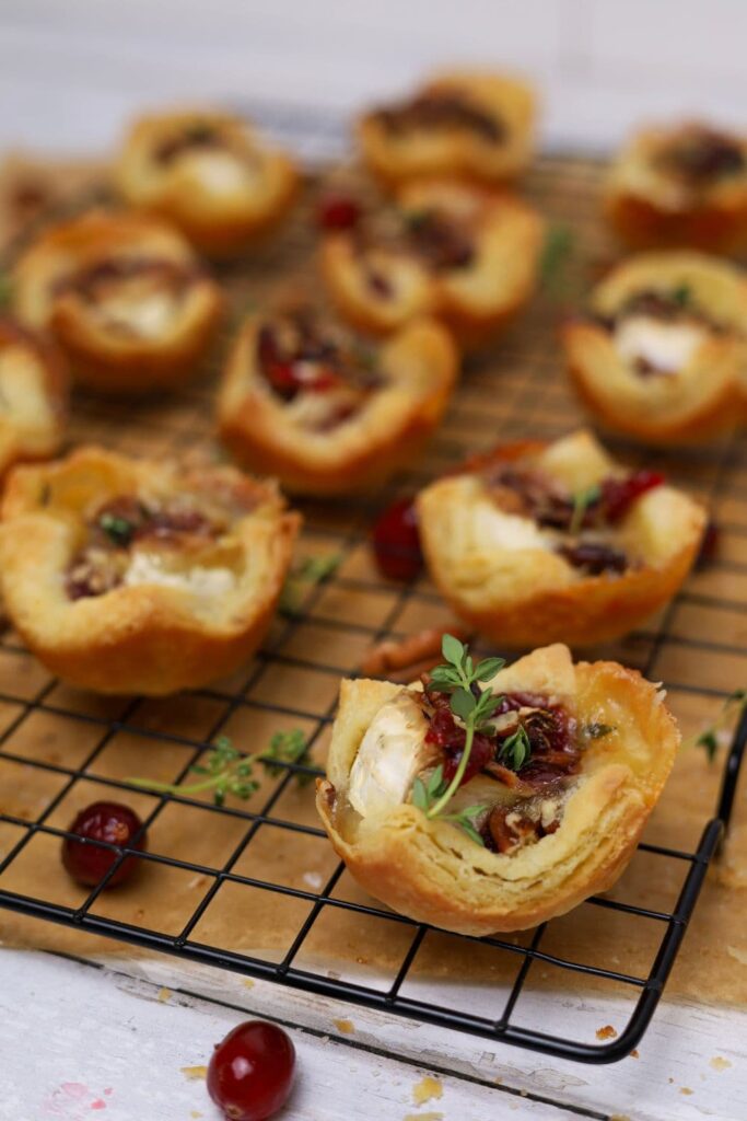 A black wire cooling rack containing 12 sourdough Brie cranberry puff pastry tarts sprinkled with pecans and fresh thyme.