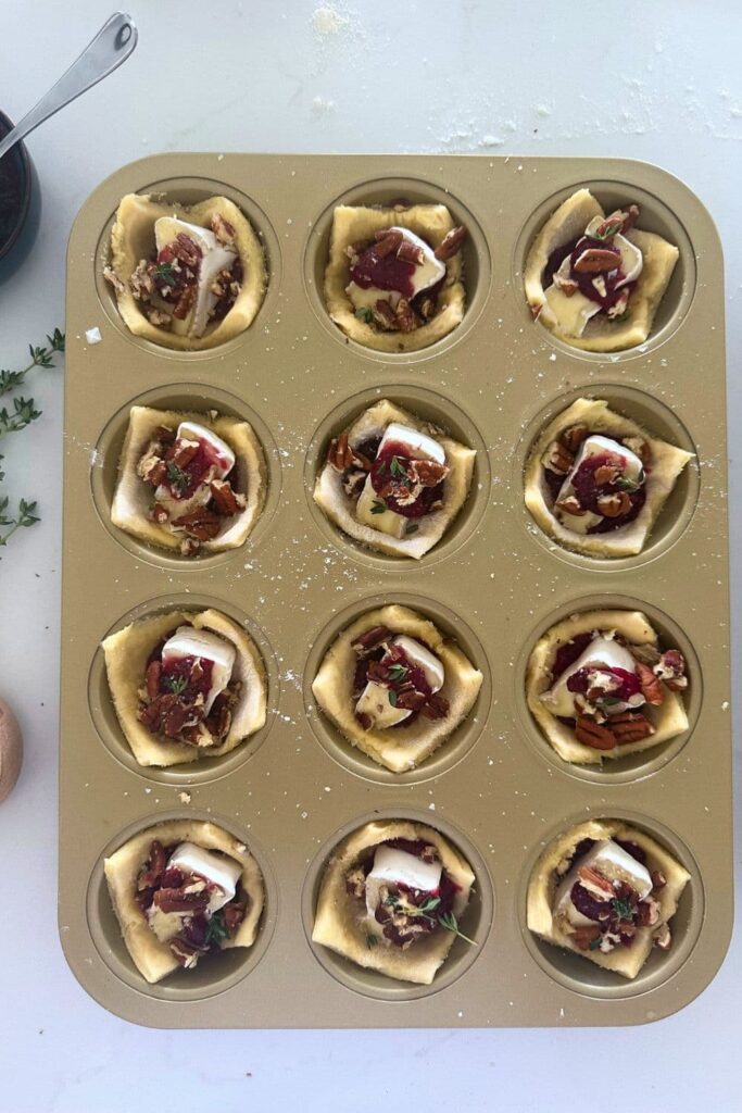 A gold muffin tray containing 12 sourdough puff pastry Brie and cranberry tarts. The tarts are unbaked.