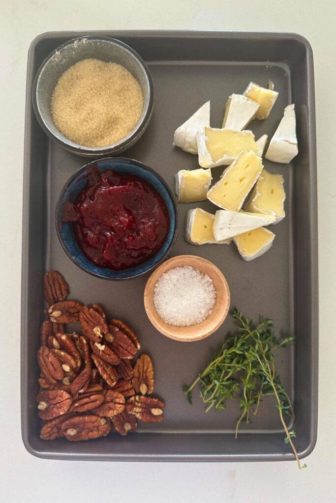 A rectangle baking tray containing small wedges of Brie cheese, pecan, cranberry jam and pecans to create small sourdough puff pastry tarts.