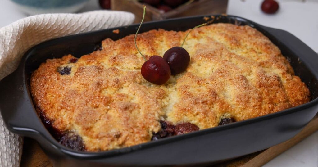 Horizontal image of a sourdough cherry cobbler that has just been removed from the oven. It has two fresh cherries placed on top.