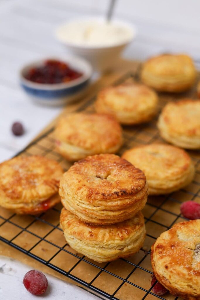 Two sourdough apple and cranberry hand pies stacked on top of each surrounded by other hand pies on a black wire cooling rack. You can see a bowl of filling in the background.