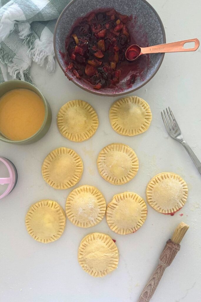 A selection of sourdough hand pies that have been filled with apple and cranberry filling sitting on a white countertop. There is a bowl of apple and cranberry filling sitting near the pies. 