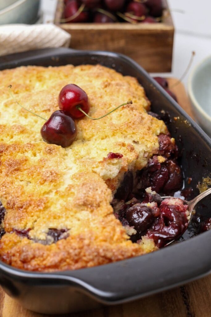 A black baking dish containing a sourdough cherry cobbler that has a big scoop taken out so you can see the glossy cherry filling underneath.