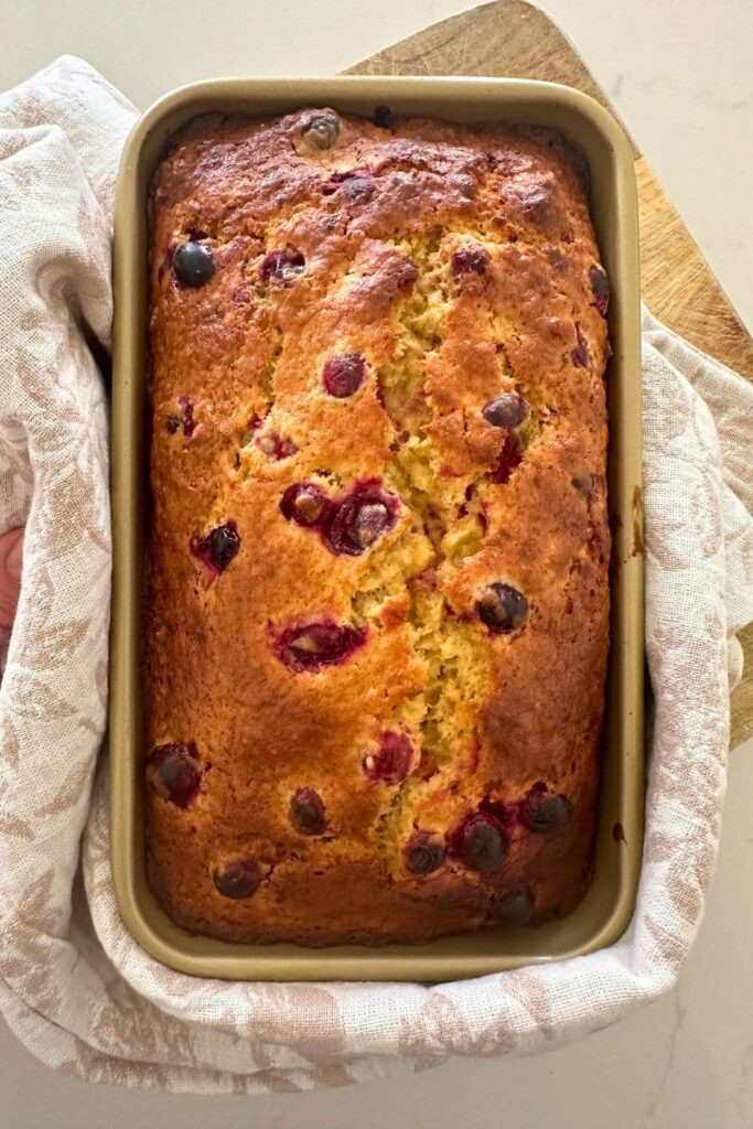 A loaf of sourdough cranberry banana bread baked in a gold loaf pan that has just been pulled out of the oven and placed onto a floral dish towel to cool. 