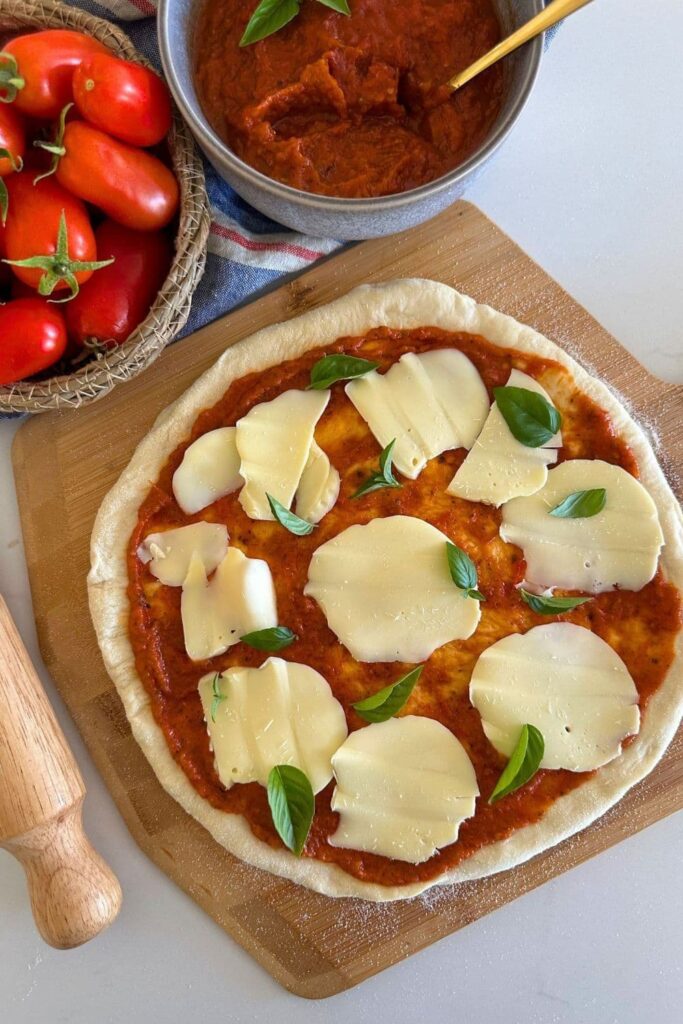 A sourdough pizza base topped with tomato pizza sauce, mozzarella cheese and basil. You can also see a bowl of roma tomatos, a bowl of homemade pizza sauce and a rolling pin the photo.