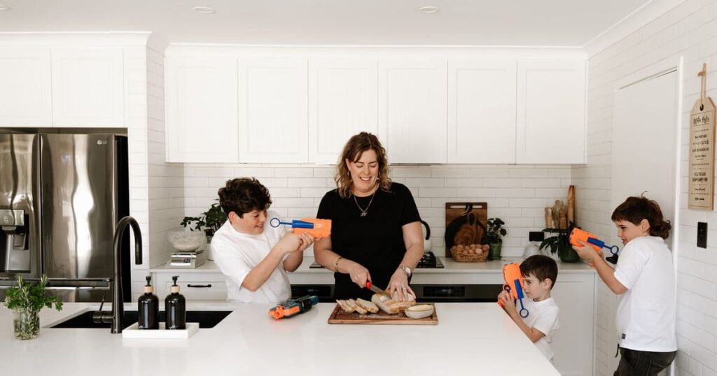 A mum in the kitchen slicing up a loaf of sourdough bread while kids play with Nerf blasters around her.