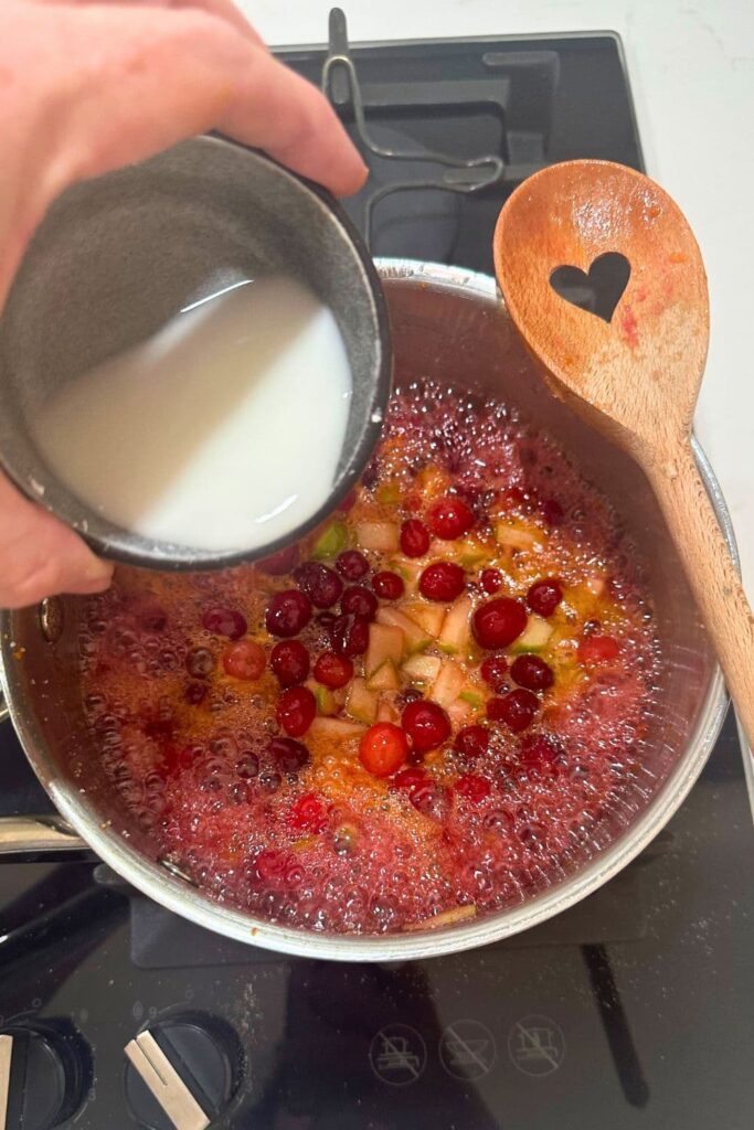 A bowl of corn starch and water being poured into the saucepan of apple and cranberry mixture that has simmered down on the stove top.