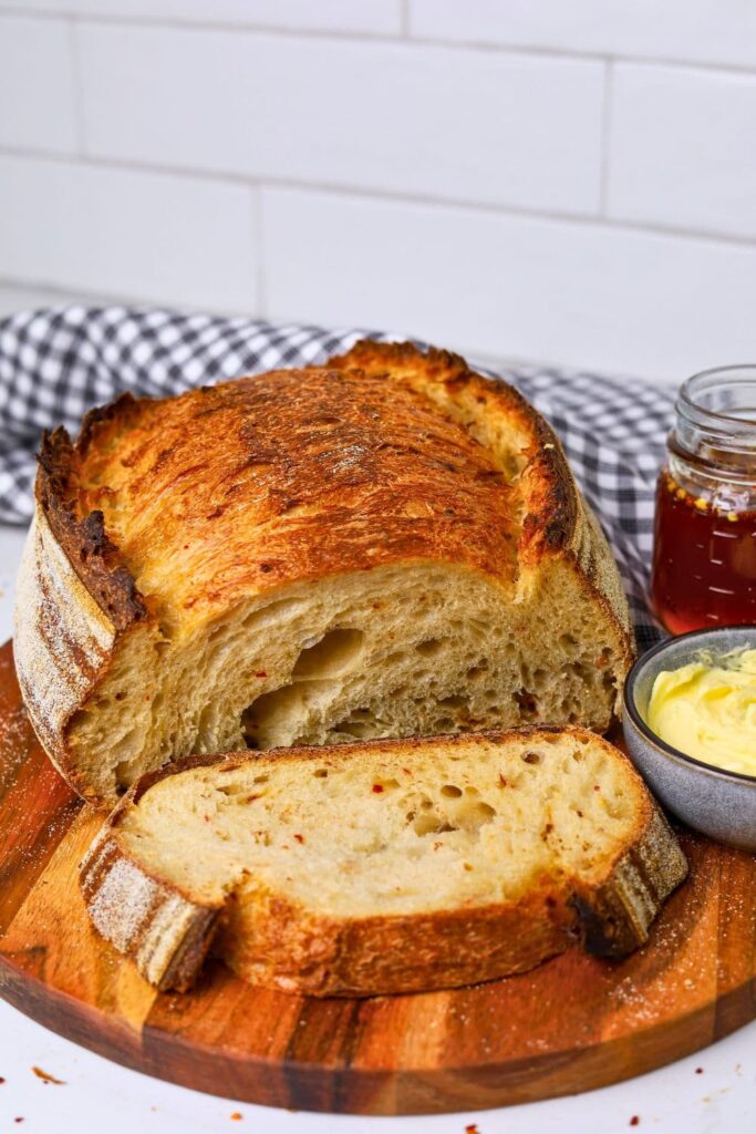 A loaf of hot honey sourdough bread that has been sliced open. You can see the crumb inside. There is a jar of hot honey and a dish of butter sitting to the right of the sourdough loaf.