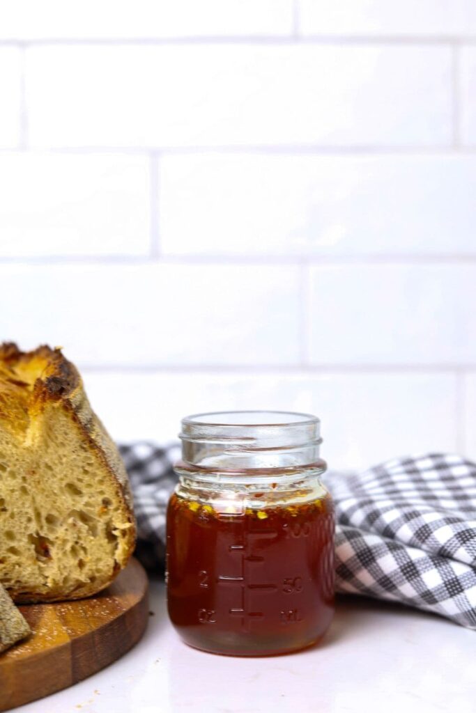 A small jar of hot honey sitting in the centre of a white tiled wall. There is a black and white checked dish towel sitting behind the jar and a loaf of hot honey sourdough bread sitting ot the left of the jar.