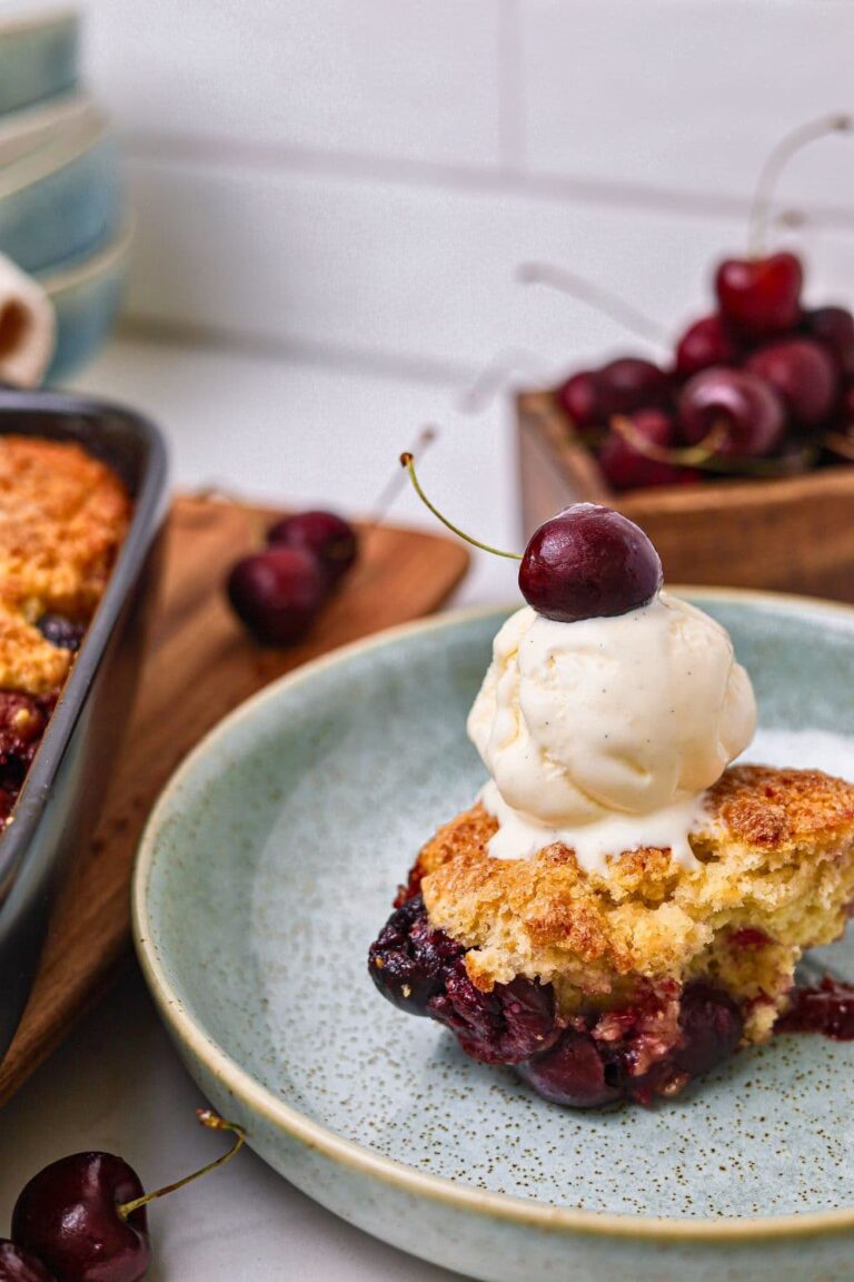 A single serving of sourdough cherry cobbler sitting in a sage green bowl. The cobbler has a scoop of vanilla ice cream on top of it and a single cherry on top.